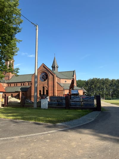 A brick church with a green roof and a steeple stands under a clear blue sky. A large tree with green leaves is on the left side of the image. A concrete utility pole with wires is in the foreground. In front of the church, there is a black metal fence, a patch of grass, and some construction materials, including an excavator and stacks of stone blocks. A sign on the stone blocks reads "SOLID SECURITY". The church is located in Stanowice, Poland.