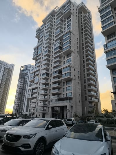 The photograph depicts a contemporary urban scene in Ashkelon, Israel, focusing on a cluster of high-rise apartment buildings. The buildings are modern in design, characterized by light-colored facades, numerous balconies with glass railings, and large windows. The architecture suggests a relatively new development. In the foreground, several white cars are parked in a lot. One car, a white Opel, is prominently featured, displaying an Israeli license plate that reads "650-19-301". The sky above is partly cloudy with hues of yellow and orange, indicating either sunrise or sunset. The overall impression is one of a residential area in a developed city. There are no people visible in the image, and no particular actions or interactions are depicted. The environment appears to be a parking area adjacent to the apartment complexes.