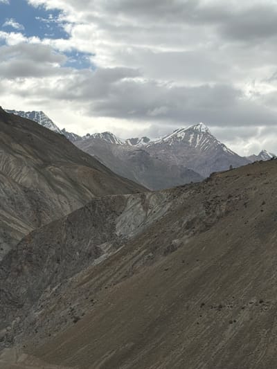 The image depicts a vast, mountainous landscape under a cloudy sky. The foreground is dominated by the rugged, barren slopes of mountains, characterized by dry earth, rocks, and visible erosion patterns. These slopes are devoid of vegetation. In the mid-ground, more mountainous terrain is visible, with a mix of rocky and scree-covered surfaces. The background features towering, snow-capped mountains, their peaks sharp and distinct against the overcast sky. The clouds are a mix of white and grey, with patches of blue sky peeking through in the upper left corner. The overall impression is one of a remote, high-altitude environment, characteristic of regions like the Himalayas in India, potentially near areas like Testa, known for its dramatic mountainous geography. There are no people or man-made structures clearly visible in the image.