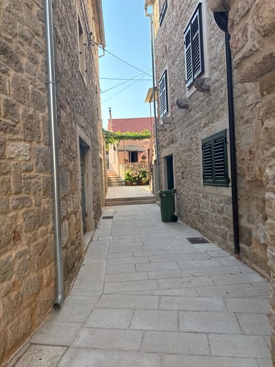 A narrow, paved alleyway winds between two old stone buildings under a clear blue sky in Jelsa, Croatia. The stone walls are constructed from irregular, roughly cut blocks. On the left, a building features a large, metallic downspout running vertically along its facade. The building on the right has windows with dark green shutters, and a dark pipe runs vertically near its edge. A green plastic garbage bin with wheels stands against the right wall. Further down the alley, the path leads towards a sunlit area where a pink building with a red-tiled roof is visible. Flowering plants in pots are arranged near the steps leading up to this building, and an umbrella provides shade. The ground is paved with rectangular light-colored stone tiles.