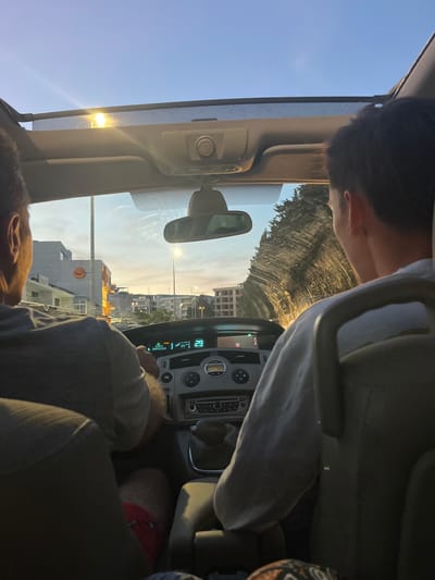 The image is a point-of-view shot from inside a car, looking forward. Two individuals are visible. In the foreground, a person is seated in the driver's seat, their back to the camera, wearing a grey shirt and red shorts. To their right, another person is in the passenger seat, also facing forward with their back to the camera, wearing a light-colored long-sleeved shirt. The car has a sunroof through which the sky is visible. The dashboard of the car is in the center of the frame, displaying various controls and a digital readout. Outside the car, a cityscape with buildings and streetlights can be seen, bathed in the warm light of either sunrise or sunset. The overall impression is of a drive through a town during twilight. The location is identified as Pistula, Montenegro.
