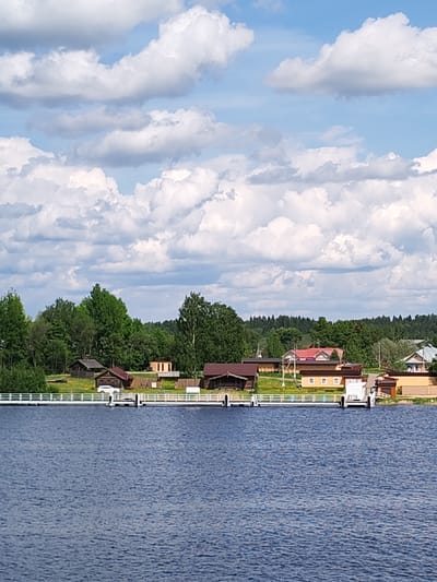The image captures a view of the Siverskoye Lake shoreline near Goritsy, Russia, characterized by a blend of natural and man-made elements under a partly cloudy sky. A body of water, possibly a lake or river, dominates the foreground with a visible surface texture indicating slight ripples. A white railing stretches horizontally across the middle ground, acting as a barrier along the shoreline. Behind the railing is a collection of buildings, a mix of cabins, houses, and structures. The rooftops vary in color, ranging from brown to red, some with visible windows. Green vegetation is abundant, including trees and bushes, adding depth to the scene. Above, the sky is filled with numerous white clouds against a light blue background.