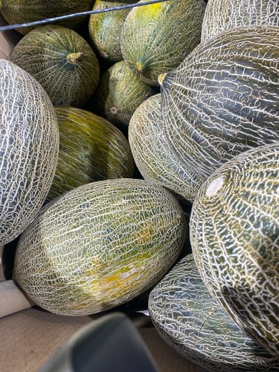 The image shows a close-up, overhead view of a pile of ripe melons, likely in a market or grocery store setting in Bel Air, Spain. The melons are characterized by their green rinds with intricate, light-colored netting patterns. Some of the melons have slight yellow or golden undertones, suggesting ripeness.
The lighting appears to be artificial, common for indoor retail environments, and there are no indicators of the time of day or weather conditions. The scene is focused entirely on the melons, showcasing their texture and natural variations. The melons are closely packed together, creating a visually dense composition. The background includes hints of wooden crates and a dark object, possibly a shelf or a piece of equipment, further reinforcing the retail environment. There is no visible text in the image. The overall impression is one of abundance and the visual appeal of fresh produce.