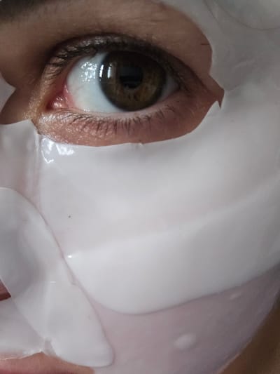 This close-up, vertically oriented media file features a person's eye. The eye is brown with a visible reflection of light on the pupil and sclera. Eyelashes frame the eye, and the skin around it appears to be slightly wrinkled. A white, sheet-like object, likely a cosmetic mask, covers a significant portion of the face, obscuring most of the features except for the eye. The mask has a smooth, slightly wet appearance and is draped in a way that highlights the contours of the face. The background is out of focus, but the overall lighting suggests an indoor setting, possibly during the day. No text is visible in the media file.