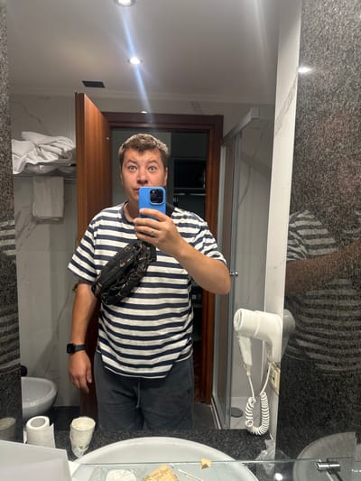 A man stands in a bathroom, taking a selfie in a mirror. He is wearing a striped t-shirt and shorts, with a fanny pack around his waist. A blue smartphone is held in his right hand, and he is looking at the camera with a surprised expression. The bathroom appears to be in a hotel room, with a shower stall, a sink, and a hairdryer mounted on the wall. The man is wearing a watch on his left wrist. The image was taken in San Martino Buon Albergo, Italy.