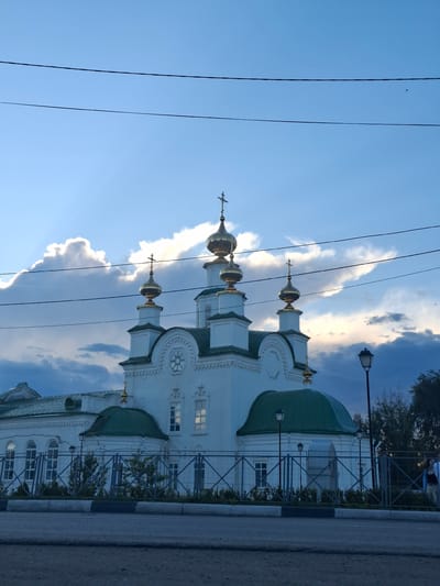 The image captures an Orthodox church in Kungur, Russia, characterized by its white walls and prominent golden domes, topped with crosses. The sky above is a dramatic blend of blue with illuminated white clouds, suggesting either early morning or late afternoon light, with sunbeams piercing through the clouds. Two dark power lines are visible stretched across the upper portion of the frame. The church is enclosed by a metal fence, behind which low bushes and trees are present. A street lamp stands to the right of the church, and a road is in the foreground. A few individuals are partially visible on the far right, near the fence, suggesting a public space. The overall impression is one of a serene and possibly historically significant religious site against a dynamic sky.