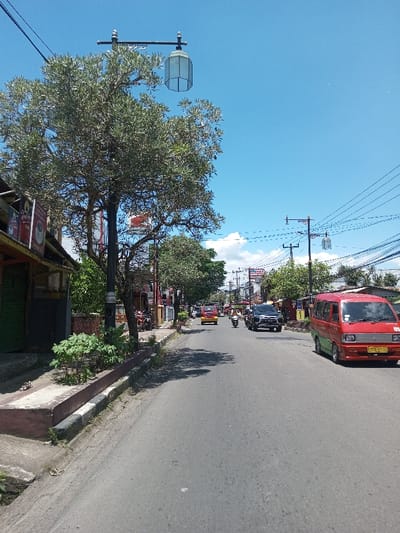 The image depicts an active urban street scene under a clear blue sky with sparse white clouds. The asphalt road, running from foreground to background, features light traffic. On the immediate right, a red and green Suzuki Carry minivan with a yellow Indonesian license plate is present, facing away from the viewer. Further down the street, a black SUV and a yellow minivan are visible, along with multiple motorcycles, all traveling in the same direction. On the left side of the road, a raised concrete sidewalk is bordered by a planter box with green foliage. A tall, dark pole with an ornate arm supports a hexagonal street lantern, partially obscured by a mature leafy tree. A row of single-story commercial buildings lines the left side, displaying varied facades and signage, including what appears to be an Indonesian flag and a sign for "RM. NUSANTARA" further down. Numerous utility poles with a dense network of overhead power lines are visible on both sides of the street. The depicted location is in the city of Tagtse Tashidingkha, Bhutan.