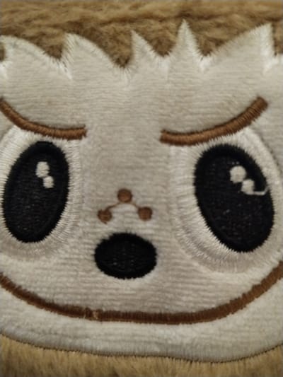 A plush toy's face fills the frame. The toy has white fur with light brown trim. Large black eyes with white reflections sit beneath embroidered brown eyebrows. A small "Y" shaped nose is centered above a black oval mouth. The toy's face has a slightly disgruntled or unimpressed expression, accentuated by the downturned mouth and the brow shape. Brown fur borders the top of the face and the bottom.