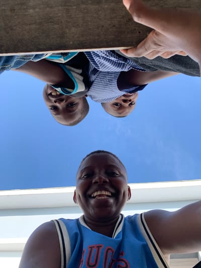 Captured from a low-angle perspective, this image shows three young boys looking down towards the camera against a clear, bright blue sky in Ibasa, Nigeria. On the right side, one boy, wearing a blue and white basketball jersey with an illegible red text, smiles broadly, revealing his teeth. On the left, a rustic wooden surface occupies the foreground, with visible hands gripping it. Behind this surface, two other boys are visible. The boy positioned higher wears a blue and white striped button-up shirt and has a neutral, curious expression. Below him, the third boy, in a striped t-shirt (blue, black, and grey) and denim, also smiles. The background is an expanse of cloudless blue sky, indicating clear, sunny daytime weather. A narrow white vertical structure is visible on the far right. The collective gaze and smiling faces suggest a joyful and engaging interaction.