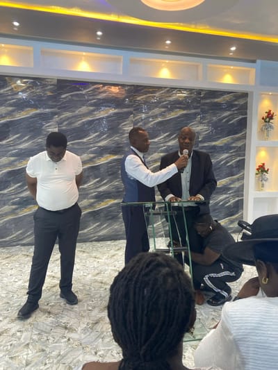 In Gbawe, Ghana, an indoor scene depicts five individuals. In the foreground, the back of a person's head with dreadlocks is visible, along with the partial view of another person wearing a black hat and white clothing to the right. Three men stand in the midground. To the left, a man in a white polo shirt, dark trousers, and black shoes stands with hands clasped behind his back, looking downwards. Central to the frame, a man in a white long-sleeve shirt, blue waistcoat, and dark trousers holds a black microphone up to a man in a dark suit jacket and light shirt, who is speaking into the microphone while standing behind a glass podium. A fourth individual is crouched on the floor behind the glass podium, wearing a dark t-shirt and dark track pants with white stripes, appearing to adjust something at the podium's base. The background features a wall covered in dark grey and white marbled tiles, with white sections above incorporating recessed shelves holding red and silver floral decorations. The floor is covered in light-colored marbled tiles. Overhead, the white ceiling displays linear and circular recessed lighting fixtures.