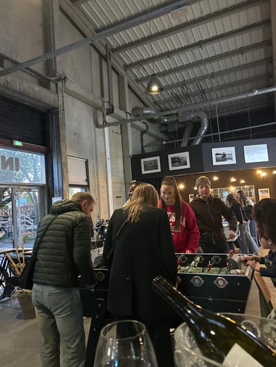 This image captures an indoor social gathering within a venue in Villeneuve-d'Ascq, France, characterized by an industrial-chic aesthetic. The high ceilings feature exposed metal beams, concrete structures, and visible ventilation ducts and cable trays. Walls are adorned with several framed black-and-white photographs and warm, industrial-style lighting fixtures, creating an inviting ambiance.
Multiple people, appearing to be adults and young adults, are present and engaged in leisure activities. The central focus is a group gathered around a black foosball table with red and blue player figures. One person in a red sweatshirt, which has partially visible text like "L.A. GELS," stands prominently near the table. Another individual in a dark jacket with blonde hair is also visible near the game. In the foreground, a dark green wine bottle and several wine glasses are visible, suggesting drinks are being served. Further into the background, a person wearing a dark green puffer jacket and light-colored jeans stands with their back largely to the camera, looking towards a large glass door or window.
The activity primarily revolves around playing or watching the foosball game and socializing. The overall mood appears relaxed and convivial. The indoor lighting is warm and ambient, suggesting evening hours, though some natural light is visible through the exterior glass door/window, where a bicycle and trees can be partially seen. Reflected on the glass, the reversed letters "МИ" are discernible.