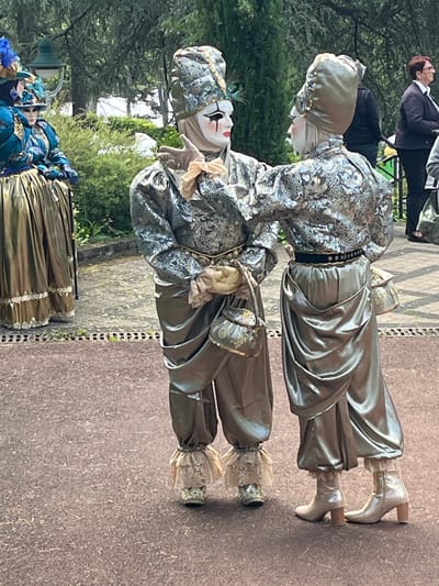 The image shows two people dressed in elaborate, old-fashioned costumes and makeup, characteristic of Venetian carnival figures. They stand close together on a paved surface, with greenery and other people in the background. The costumes feature silver and grey fabrics, lace details, and heeled boots. One figure holds a small purse. The scene appears to be outdoors, possibly a public space with trees and other decorative figures in the distance. Based on the context provided, it is plausible that the location is Saint-Galmier, France.