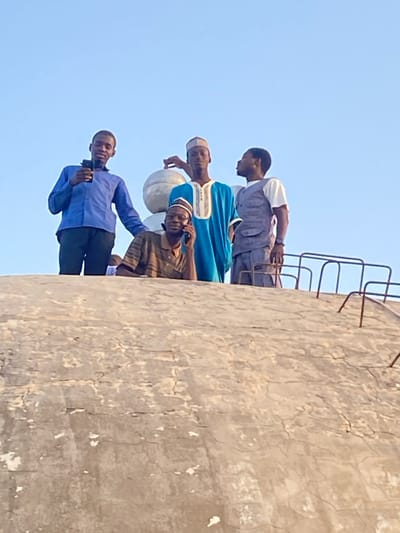 Four males are observed atop a large, light grey concrete dome or roof structure in Barako, Nigeria, against a clear blue sky.
The concrete surface, showing signs of weathering and minor cracks, occupies the lower half of the frame. A dark, rusted metal railing, possibly part of an access ladder or safety barrier, is visible on the right side of the concrete. Behind the standing figures, two metallic, light-colored spherical objects, possibly architectural finials, are present.
From left to right:
1. A young adult male, dressed in a blue long-sleeved shirt and dark trousers, stands with his left hand holding a black smartphone vertically, directed slightly upwards. His right arm is extended towards the adjacent individual.
2. A young adult male stands centrally, wearing a light blue traditional tunic with white embroidery and a light-colored cap. His right hand rests on one of the spherical objects behind him. He is looking towards the right.
3. An older adult male is positioned in the foreground, partially obscured by the concrete structure. He wears a brown and tan striped short-sleeved shirt and a light-colored cap. He holds a black smartphone to his right ear, actively engaged in a call.
4. A young adult male stands on the right, clad in a white short-sleeved shirt, a grey vest, and grey trousers. His left arm is bent, and his right hand rests on the metal railing. He is looking towards the left, appearing to interact with the central standing male.