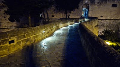 The image depicts a curved stone walkway at night, likely part of a historic fortification or castle in Taranto, Italy. The walkway is illuminated by a series of small, modern lights embedded in the low stone wall on the left, casting bright, sharp pools of light onto the wet cobblestone pavement. The stone wall itself is constructed from rough-hewn blocks, some showing signs of age and weathering.
Beyond the walkway, a larger stone wall forms the backdrop, with a dimly lit arched entrance visible in the distance, suggesting a passage into another part of the structure. Trees with dark, silhouetted foliage are visible against the stone walls, adding to the nighttime atmosphere. The cobblestones are wet, reflecting the lights and creating a shimmering, almost ethereal effect, hinting at recent rain or dampness. The overall mood is mysterious and atmospheric, emphasizing the age and grandeur of the location under the cloak of night. No people or text are visible in the image. The time of day is clearly night, and the weather appears to be damp.