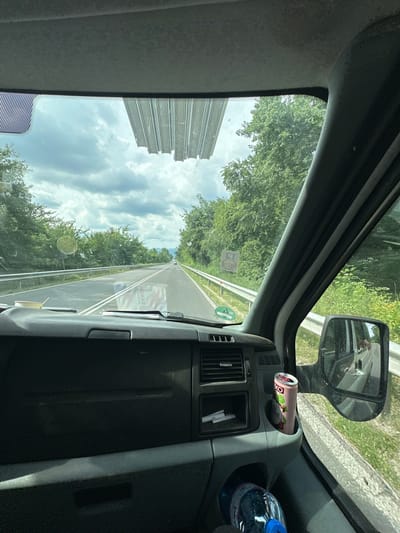 The image is taken from inside a vehicle, likely a truck, looking out through the windshield and side window. The scene is a highway with trees lining both sides. The road appears to be a two-lane highway with white markings. The sky is cloudy, indicating a day with mixed weather.
Inside the vehicle, on the dashboard, a can of what appears to be a beverage is placed in a holder. To the right, a plastic bottle of water with a blue cap is visible. A side mirror reflects the road and sky. There is a gray interior of the vehicle visible, including part of the dashboard and a vent.
Outside, a tall, gray, rectangular structure is visible to the left, possibly a building or industrial structure. The trees are lush and green, suggesting a warmer season. There are two stickers visible on the outside of the vehicle's window, one with a circular design and the other a green circular sticker with a white symbol and a green outline.
The time of day appears to be daytime, given the lighting. The overall impression is of a journey along a highway on a somewhat overcast day. No people are clearly visible inside the vehicle, although a reflection in the side mirror might indicate a person. The image is oriented vertically, making the view appear as if the vehicle is driving upside down.