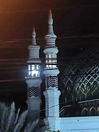 The image shows two minarets and the dome of a mosque at night. The minarets are white with black chevron patterns and tiered sections. The dome is green with a geometric metal framework. The surrounding environment is dark, with visible streetlights and utility wires. Palm trees are present in the foreground. The architecture and style are consistent with mosques found in Indonesia. It is located in the city of RawangItiek, Indonesia.
