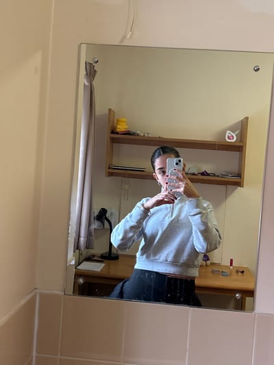 The image displays a mirror selfie captured by a female individual within an interior room, likely student accommodation at Edge Hill University in Ormskirk, West Lancashire, United Kingdom. The individual is reflected from approximately the waist up, wearing a light grey, long-sleeved, quarter-zip cropped sweatshirt and dark lower garments. Her dark hair is pulled back, and her fingernails are painted dark red. She is holding a white smartphone horizontally with both hands, positioned to capture her reflection.

The large rectangular mirror is affixed to a light-colored wall, which has a lower section of light beige/pinkish tiles. Reflected beneath the mirror is a wooden desk. On the desk, a black desk lamp with a flexible neck is visible on the left, resting near white papers. To the right on the desk, a red tube (possibly lipstick) and two small, circular containers are present. Above the desk, two wooden shelves are mounted. The top shelf holds several yellow sponges or similar items, miscellaneous toiletries, and a white object with a pink interior. The lower shelf contains books or notebooks on the left, and additional indistinct items on the right. A small, light-colored figure hangs below the lower shelf. A light-colored curtain is visible reflected to the left of the desk lamp, and a white cable descends from the upper wall in the reflection. The room exhibits neutral-colored walls, consistent with a functional living space.