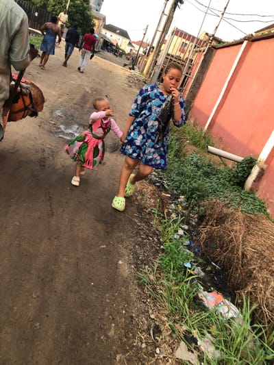 Two children stand on a street near a building and overgrown foliage. The older child wears a blue patterned dress and light green shoes. The younger child, close by, is in a pink and green patterned dress. Several adults walk in the background. In the foreground, part of an animal-drawn cart is visible, with what appears to be a drum and a person in a light green garment. The ground is unpaved.