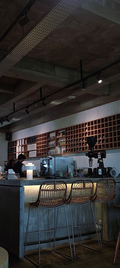 The image depicts the interior of a modern cafe in Cicalengka, Indonesia. The scene is dominated by a concrete ceiling with exposed conduits and track lighting, and a wooden slatted shelf unit filled with various bottles and packages. In the foreground, three rattan bar stools with metal legs are positioned in front of a concrete counter. Behind the counter, a barista, a person wearing a dark vest over a white shirt, is interacting with a customer. A bright lamp illuminates the counter area. Various cafe equipment, including a coffee grinder and an oven, are visible behind the counter. The overall aesthetic is industrial yet cozy, with warm lighting highlighting the textures of the materials.
