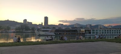 The image depicts a sunset scene along the Danube River in Linz, Austria. In the foreground, several individuals are seated on the grassy riverbank, some facing the water, while others are turned away from the camera. A paved path is visible in the lower portion of the image.
Dominating the middle ground is a large, white river cruise ship named "THE GENTLEMAN," docked on the river. The ship features multiple decks, with rows of windows and lounge chairs visible on the upper levels. The ship's name is illuminated on its side.
The background reveals the cityscape of Linz, with various buildings, including modern high-rises and older structures, set against a backdrop of rolling hills. The sky is a gradient of warm yellow and orange hues from the setting sun, with some clouds present. The river reflects the colors of the sky and the lights of the city. The overall atmosphere is tranquil and serene, capturing a moment of relaxation by the river at dusk.