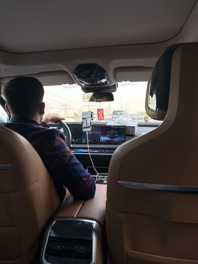 Inside a car, a man sits behind the steering wheel, facing forward. His hands grip the wheel, and his head is turned slightly toward the front. A large display screen dominates the center console, displaying car information. A phone is mounted above the screen. The car's interior features light brown leather seats, a cream roof, and metallic trim. Out the front window, other cars and buildings are visible.
