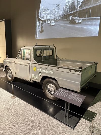 The image displays a vintage light grey Datsun G222 Model truck on exhibit in an indoor setting, likely a museum or exhibition hall in Sumida, Japan. The truck, a compact utility vehicle, is positioned on a dark, raised platform, viewed from its left side and slightly from the front. On the side of the truck bed, "NAKAJIMA-SHOTEN" is painted in white capital letters, accompanied by the Japanese characters "自家用" (Jiyouyou - for private use) and a yellow circular emblem.
To the left of the truck, a large black and white image is projected onto a wall, depicting an urban street scene with buildings and what appear to be other vehicles, providing historical context for the truck's era or typical environment. To the right of the truck, a black information placard with white text is visible. The placard is titled "Datsun Truck G222 Model" in both Japanese and English. The English text explains that the truck is "one of the 220 model series, representing the second generation of the Datsun Truck." It further states that the "NAKAJIMA-SHOTEN" lettering was "applied at the time of purchase" by a father who operated a packing business, and the vehicle was used until 2007, preserving the "atmosphere of a Showa 30s (1955-1964) vehicle." The floor of the exhibition space is covered with a light-colored, patterned carpet. The scene presents a well-preserved historical vehicle in an informative display setting.