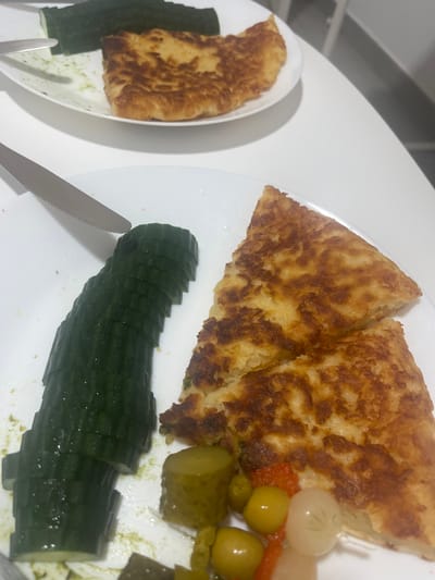 A high-angle, indoor shot depicts a plate of food on a white surface. The plate contains two triangular slices of fried bread, a sliced cucumber, and a variety of pickled vegetables including olives, pickles, and pickled onions. A knife rests adjacent to the sliced cucumber. A second plate with a smaller piece of fried bread and sliced cucumber is visible in the upper-left corner. No people are visible. The location is implied to be within Mogán, Spain based on the user prompt.