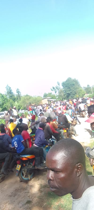 The scene is a crowded, dusty dirt road in Isukha ICHINA, Kenya, with a large group of people congregated. Many people are riding motorcycles and there are several parked in the background. Some people are standing and talking, while others are walking. There are some green trees in the background, and a bright blue sky above. One man in the foreground is wearing a grey shirt and a mustache. He is looking to the left side of the image. This is likely a scene from a local market or festival.