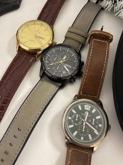 The image displays three wristwatches arranged diagonally on a white, speckled surface, suggesting an indoor setting. The top watch has a gold-toned case and a patterned brown leather strap. Its dial is gold with black hour markers and a date window. The middle watch features a black case with a gray leather strap and a black dial with yellow accents and sub-dials. The bottom watch has a silver-toned case, a brown leather strap with white stitching, and a dark gray dial with white hour markers and sub-dials, one of which prominently displays the number '12'. The watches appear to be new or in good condition, and there are no visible people or animals in the scene. The context of Slobozia-Dușca, Moldova, is not directly represented by any visual cues within the provided image.