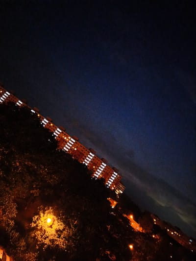 The image depicts a nighttime view of apartment buildings in the city of Most, Czechia. The buildings are illuminated with numerous lit windows, creating a grid-like pattern against the dark sky. The scene is angled, with the buildings running diagonally from the lower left to the upper right of the frame. In the foreground, there are trees and vegetation, some of which are illuminated by streetlights, casting a warm, orange glow. The sky is a deep, dark blue, with subtle variations in shade and some visible noise or grain in the image. The overall impression is that of a residential area at night.