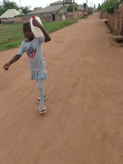 A young girl with braided hair walks down a dirt road in Barakin Angundi, Nigeria, carrying a large white plastic jug with a red lid. She is wearing a grey t-shirt with a graphic design and the words "Girl Power" printed on it, along with grey leggings and sandals. The road is lined with buildings and vegetation. In the background, other people can be seen walking further down the road. The sky is overcast.
