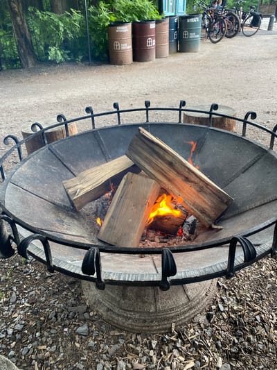 The image shows a fire pit with burning logs and embers. The fire pit is large and made of metal with a decorative wrought iron surround. It is set on a stone base. In the background, to the left, there are several bicycles parked in a row. There are also some cylindrical barrels with text and symbols on them, which appear to be recycling bins. Green foliage is visible to the far left. The ground around the fire pit is covered with gravel and small stones. The scene suggests an outdoor setting, possibly a park, campsite, or an outdoor recreational area. The presence of bicycles and recycling bins might indicate a public space. The lighting suggests it could be daytime, with the fire providing a warm glow. The overall atmosphere appears relaxed and inviting, with the fire suggesting warmth and a gathering place.

There is visible text on the barrels. One barrel has "БУМАГА" and "ПЛЭЛ ПОРТОЯН" printed on it. Another barrel has "ОБЩЕ ОТХОДЫ" and a symbol of a house with a window. Another barrel has "СТ" and a symbol of a house with a window. Another barrel has "АЛЮ" and a symbol of a house with a window. Another barrel has a symbol of a house with a window.
