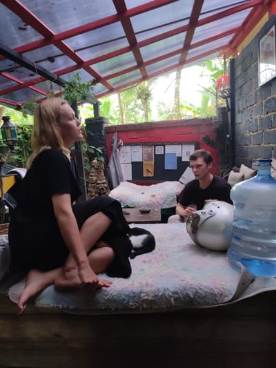 The scene is set in what appears to be an outdoor lounge area in Bedulu, Indonesia. A woman with blonde hair, wearing a black dress, is seated with her legs crossed. Her feet are bare. Next to her is a black and white cat. A man is seated to the right, wearing a dark t-shirt. In front of him is a white motorcycle helmet and a large bottle of water. The area is under a translucent roof with red supports, and there is greenery visible through the open walls, along with a brick wall, a painted red wall, and informational postings. The setting evokes a casual and relaxed atmosphere.