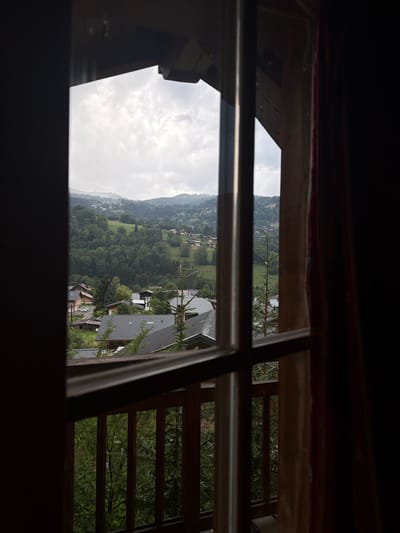 The image is a photograph taken from a window or balcony, looking out onto a scenic landscape. The foreground is dominated by dark, silhouetted wooden window frames and a metallic railing, suggesting an interior viewpoint. Through these frames, a picturesque view of a village nestled in a valley unfolds.
The scene is a hillside village with numerous houses and buildings with dark, pitched roofs, interspersed with lush green trees and rolling hills. The architecture appears to be traditional, with some buildings having wooden balconies and structures. A road or path winds through the village, and the density of the trees suggests a rural or mountainous setting. The sky is overcast with a soft, diffused light, indicating it might be morning or late afternoon, and the weather appears to be cloudy.
In the distance, the hills recede into a hazy, bluish-grey backdrop, creating a sense of depth and vastness. The overall atmosphere is tranquil and serene, conveying a sense of peaceful coexistence between human habitation and nature. The image captures a moment of quiet observation, inviting the viewer to contemplate the beauty of the landscape. There is no visible text in the image.