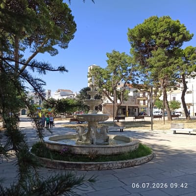 In Ruvo di Puglia, Italy, a sunlit public square features a three-tiered fountain with equine sculptures at its base. The fountain is surrounded by grass and situated on a paved area. Benches are scattered throughout the square, some occupied by people. Tall pine trees with lush green foliage frame the scene, casting shadows on the ground. In the background, buildings, including a tower with arched windows, and a playground are visible under a clear blue sky. The timestamp "06.07.2026 09:42" is present in the lower right corner.
