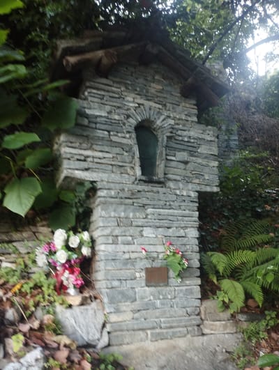 The image shows a small, rustic stone structure, resembling a shrine or a votive monument, situated in a natural, wooded environment. The structure is built from rough-hewn, greyish stones of varying sizes, stacked horizontally. It features a small, arched opening near the top, with a dark interior. Above this opening, there is a roof-like structure made of rough logs.
At the base of the shrine, and slightly to the left, a bouquet of artificial flowers, predominantly white and pink with some red accents, is placed in a container. The flowers are vibrant and appear relatively new. Further to the left, there are large, green leaves of a climbing plant, and fallen leaves on the ground.
To the right of the shrine, a cluster of green ferns is visible, with their fronds extending upwards and outwards. The overall environment suggests a slightly overgrown, natural setting, likely outdoors. The lighting appears somewhat diffused, indicating it might be overcast or the sun is not directly shining on the structure.
The shrine's construction and the surrounding vegetation suggest it is located in a natural or semi-natural area, possibly along a path or in a garden. Given the context of Orselina, Switzerland, such shrines are often found in rural or mountainous areas, possibly associated with religious or historical sites. The image does not contain any people, nor any immediate signs of human activity other than the shrine itself and the flower offering.