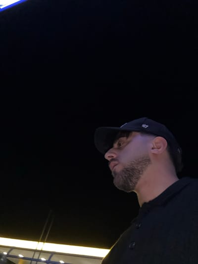 A man in a baseball cap and black button-down shirt is viewed from a low angle against a black background. He has a trimmed beard and appears to be looking down. A bright vertical light fixture is visible in the background on the right. The image is dark, with the man's face and upper body illuminated.