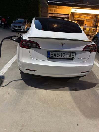 The image shows the rear of a white Tesla Model 3 electric vehicle parked in a charging bay. A charging cable is plugged into the vehicle's charging port. The Tesla has a Bulgarian license plate that reads "EA 5121 AE". In the background, there is a grey Peugeot car and a red motorcycle parked. Behind the cars, there is a building with a bar or restaurant area with lights and seating. The scene appears to be an outdoor parking lot with a concrete surface and white painted lines indicating parking spaces. The time of day appears to be evening or night, given the lighting. It is not possible to identify specific details of Edirne Merkez, Turkey from this image.