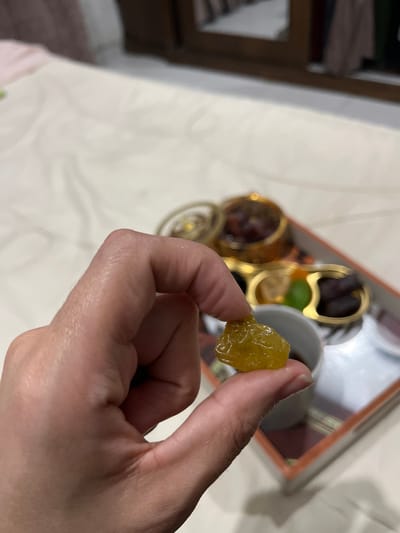 A person's hand, seen from a slightly elevated perspective, holds a single, translucent, yellow, irregularly shaped object—possibly a candied fruit or large golden raisin—between the thumb and index finger. The object appears moist or sticky. In the soft-focus background, a rectangular tray with an orange-brown rim rests on a light-colored, textured surface, possibly a bedspread. On the tray, a small white cup contains a dark liquid, alongside an ornate golden lidded container and a segmented golden dish holding an assortment of items: dark-colored dates, lighter dried fruits, and a single green piece. Distant background elements include blurry, dark-toned furniture. The scene is an indoor setting in Nuseirat, Palestinian Territory.
