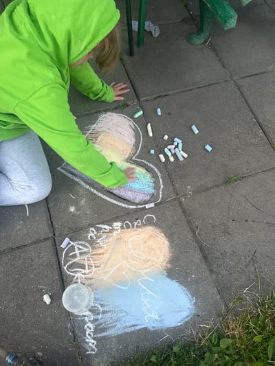A child wearing a bright green hoodie and gray pants is drawing with chalk on a paved surface, likely a sidewalk or patio. The child is positioned in the lower left quadrant of the image, facing away from the camera and towards the right. Their left hand is drawing on a large chalk heart, which is filled with rainbow colors. Scattered around the drawing area are several pieces of chalk in various pastel colors.
To the right of the heart, there are two larger chalk drawings. One appears to be a large, pale orange heart next to a light blue, heart-shaped area. White chalk writing is visible within these shapes, and some symbols or drawings are present below them, including what looks like "ATT" inside a circle. Adjacent to these drawings is a clear plastic container, possibly for water or holding chalk.
The ground is made of rectangular concrete pavers, with some grass and weeds growing in the cracks and along the edges of the paved area, particularly on the right side of the image where the grass is denser and greener. A green metal bench is partially visible in the upper left corner, and a gray sock lies on the ground near it. The scene suggests a daytime activity, possibly outdoors given the presence of grass and the informal drawing on the ground. The child's focus on their artwork and the colorful chalk drawings evoke a sense of creativity and playfulness. The visible writing includes Cyrillic letters, with "семья" (family) being discernible on the blue chalk heart.