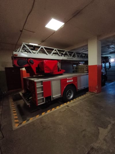 In a dimly lit indoor structure, possibly a fire station bay in Tukums, Latvia, a red and silver fire truck with a deployed ladder is parked. The truck features the "MAGIRUS" brand name visible on the ladder. The environment is utilitarian, with a concrete floor marked by yellow and black hazard stripes. A white support column, painted red in its lower section, stands to the right of the truck. Above, electrical conduits run along the textured ceiling, illuminated by a bright, square light fixture. The overall impression is one of functional storage for emergency equipment.