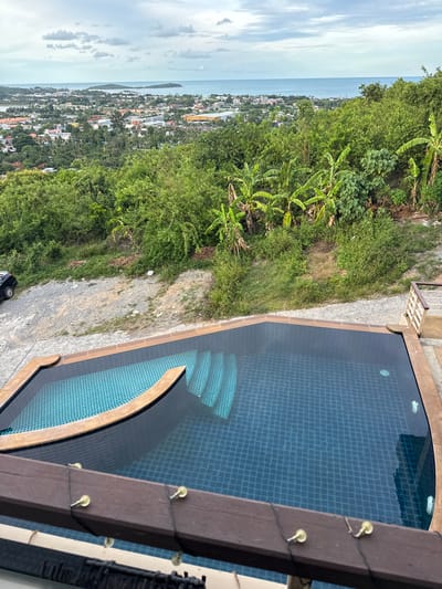 An aerial perspective captures an infinity pool with a dark blue tiled bottom and a light blue tiled shallow end, set against a lush, green tropical landscape overlooking a coastal town. Banana trees are visible amongst the dense foliage. In the distance, the ocean stretches to the horizon, meeting a cloudy sky. The town, featuring a mix of buildings with red and white roofs, is situated between the greenery and the sea. A dark-colored car is parked on a gravel lot near the edge of the vegetation. A wooden railing in the foreground frames the view. The image appears to have been taken in the late afternoon, given the soft lighting.