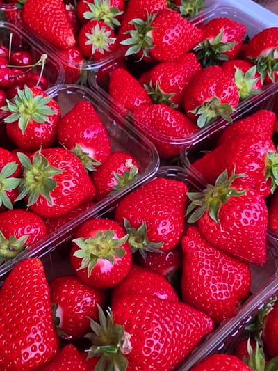 The image shows a close-up overhead view of multiple plastic containers filled with fresh, ripe strawberries. The strawberries are a vibrant red color, with visible seeds and green leafy tops. Some containers also appear to contain cherries. The scene is brightly lit, highlighting the texture and glossiness of the fruit. The containers are transparent, allowing a view of the stacked fruit within. The image appears to be taken at a market stall or a produce stand in Voluntari, Romania.
