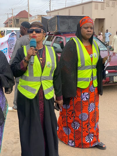 The image depicts a street scene in Rigachikun, Nigeria, where two women are central figures. The woman on the left, wearing a black dress, a black beret, and sunglasses, holds a microphone and is speaking. She is also wearing a bright yellow reflective vest over her dress, with a logo on it. The woman on the right, dressed in a black headscarf and a black open-fronted garment over an orange and white patterned dress, also wears a yellow reflective vest. She holds some papers in her hand. Both women are positioned in the foreground, facing towards the camera and appearing to be addressing an audience or participating in an event.
Behind them, a red vehicle is visible with a large speaker system mounted on top. Other people can be seen in the background, some in casual attire, and others wearing light blue and cream-colored clothing, suggesting a public gathering or procession. The environment appears to be an urban street with buildings and overhead power lines visible. The overall impression is of a public address or a community event taking place outdoors.