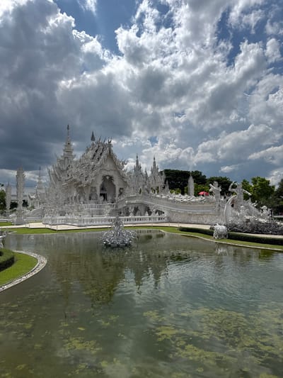 The image captures a dramatic view of the White Temple (Wat Rong Khun) in Ban Rong Khun, Thailand. The temple, a stunning example of contemporary Buddhist art, is predominantly white and features intricate, ornate architectural details. It is surrounded by a body of water, likely a pond or canal, which reflects the temple's brilliant white structure and the surrounding greenery. The water contains patches of algae and at least one visible fish, adding to the serene yet lively atmosphere.
The sky above is filled with a dynamic array of clouds, ranging from bright, fluffy white cumulus to darker, more brooding storm clouds, suggesting a changeable weather pattern, possibly late afternoon. The sunlight illuminates the temple brilliantly, highlighting its intricate sculptures and spires, while casting some shadows that add depth to the scene. Green trees frame the temple and its reflection, providing a natural contrast to the man-made masterpiece.
The temple's elaborate design includes numerous statues and sculptures, many of which appear to be mythical creatures and Buddhist figures, all rendered in white. A prominent feature is the bridge leading to the temple, adorned with countless hands reaching out from below, symbolizing the suffering of the dead. A bright red flag is visible atop one of the temple's structures.
The overall mood of the image is one of awe and wonder, emphasizing the ethereal beauty and unique artistic vision of the White Temple. The composition, with the temple dominating the frame and its reflection in the water, creates a sense of grandeur and spiritual significance. The image appears to be taken from a slightly elevated angle, offering a panoramic view of the temple complex and its immediate surroundings. No visible text is present in the image.