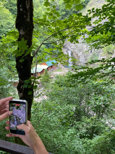 The media file depicts a person holding a smartphone, with their hands in the foreground, as they record a scenic view. The phone screen shows a live recording of lush green foliage and what appears to be a mountainous landscape.
The overall scene is a natural environment, characterized by abundant green trees and foliage, suggesting a forest or mountainous region. In the mid-ground, there is a glimpse of a waterfall cascading down a rocky cliff face into a pool of water. Below the waterfall, a cluster of buildings with colorful roofs (green, blue, and yellow) are visible, appearing to be part of a small settlement or resort.
The image is taken from a high vantage point, looking down into the valley where the waterfall and buildings are located. The presence of a phone recording the scene implies the person is experiencing and documenting the natural beauty of the location. The time of day appears to be daytime, with good natural lighting. The weather seems pleasant, conducive to outdoor activities and sightseeing. The lush greenery suggests a temperate climate, possibly during spring or summer.
The context provided places the scene in Khostinskiy, Russia, which aligns with the mountainous and forested landscape. The emotions conveyed are those of appreciation for nature and the enjoyment of a travel or leisure experience. There is no visible text in the image.