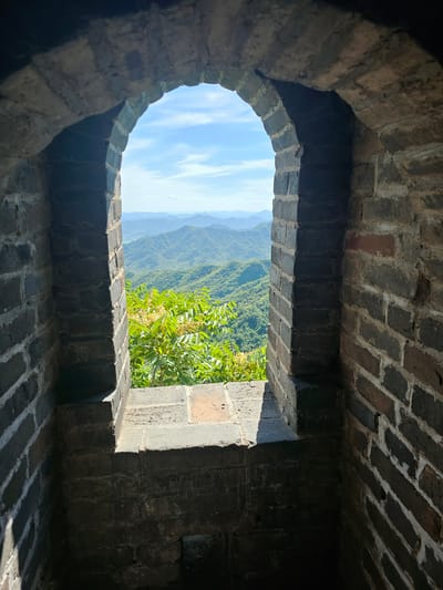 The image shows a view through an arched opening in a brick wall, likely part of the Great Wall of China, in Huairou District, China. The opening frames a landscape of lush green mountains under a bright blue sky with scattered white clouds. In the foreground, a section of the thick, dark brick wall is visible on both sides, with the archway constructed of smaller, lighter-colored bricks. A horizontal ledge made of brick runs along the bottom of the opening, casting a shadow into the interior of the structure. In the middle ground, just beyond the opening, vibrant green foliage with small yellow flowers is prominent. The distant mountains recede in layers, becoming lighter and bluer with distance, indicating atmospheric perspective. The scene is bathed in sunlight, creating a sharp contrast between the shadowed interior of the wall and the brightly lit exterior landscape.