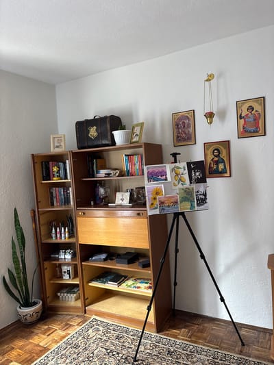 The image depicts an indoor domestic space within the Vrnjacka Banja Municipality, Serbia. The scene is dominated by a two-section wooden bookshelf unit positioned against a white wall.
On the left side of the unit, a tall, potted snake plant (Sansevieria trifasciata) rests on the parquet floor in a white decorative pot. The left bookshelf section contains multiple shelves displaying books and various art supplies, including bottles of paint, brushes in a holder, and organized containers of markers or similar items.
The right bookshelf section features a dark brown trunk adorned with a gold Hogwarts crest on its top surface. Below this, shelves hold books, a small white plant pot, and a decorative framed picture. A central pull-out or desk section of lighter wood is visible, with lower shelves containing notebooks, art supplies, and dark boxes.
The white wall above and to the right of the bookshelf unit is decorated with several framed Orthodox Christian icons: a depiction of the Theotokos with Child on the upper left, an icon of a saint (possibly a warrior saint) on the upper right, and an icon of Jesus Christ Pantocrator below and to the left of the warrior saint icon. A brass or gold-colored hanging vigil lamp (kandilo) with a red insert is suspended from the wall between the two upper icons.
In the foreground, a black tripod easel displays seven small pieces of artwork clipped to its surface. These include landscapes (mountains, palm trees), still life (lemons, sunflowers, a vase of flowers), and silhouette art (a jazz musician, an elephant). A patterned beige and brown area rug covers a portion of the wooden parquet floor in the lower center. No individuals are present in the scene.