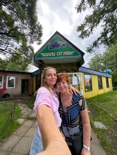 Two women are posing for a selfie in front of a yellow building. The woman on the left is wearing a pink striped shirt and blue jeans, while the woman on the right is wearing a black and blue striped shirt. Both women are smiling. The building behind them appears to be a cafe or restaurant, with a sign in Russian that reads "Вопли от жен". The building has a green roof with a smaller green sign that has a picture of a bus and the words "Счастливого пути". The surroundings are overgrown with grass and trees. The weather appears to be cloudy.