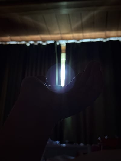 A close-up, silhouette shot from a low angle shows a person's cupped hands extended towards a bright vertical light source. The light appears to be shining through a gap in what might be curtains or blinds, creating a lens flare effect on the left side of the frame. The background is dark, with a textured ceiling or overhang visible at the top. The scene is indoors, and no specific location in Vilyuysk, Russia, can be determined from the image.